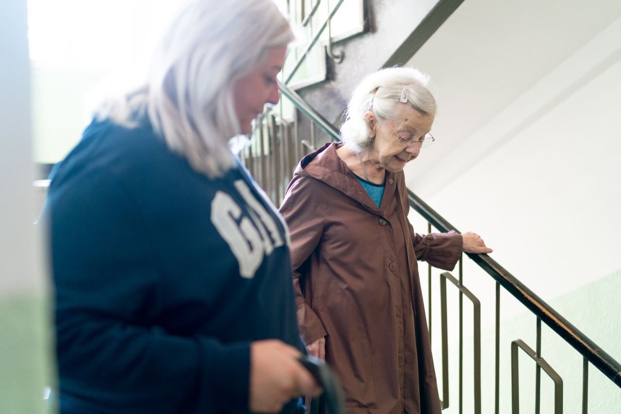 Elderly woman with caregiver on stairs, symbolizing home care and support, in Prague setting.